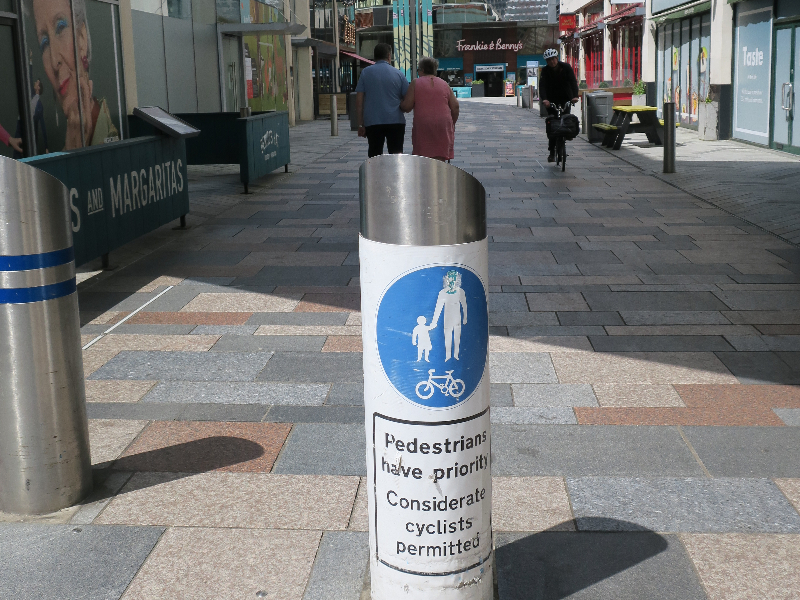 A bollard at the entrance to an external shopping area with a shared use sign with the wording below 'Pedestrians have priority - Considerate cyclists permitted'.