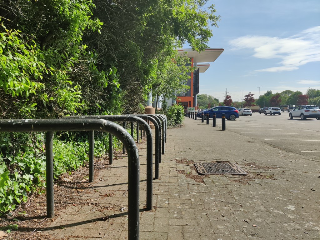 A paved sidewalk with spacious and well secured metal bike racks in the foreground, bordered by lush green vegetation on the left. To the right is a parking lot with several cars. In the background, a modern building with orange accents is visible. The sky is mostly clear with a few scattered clouds.