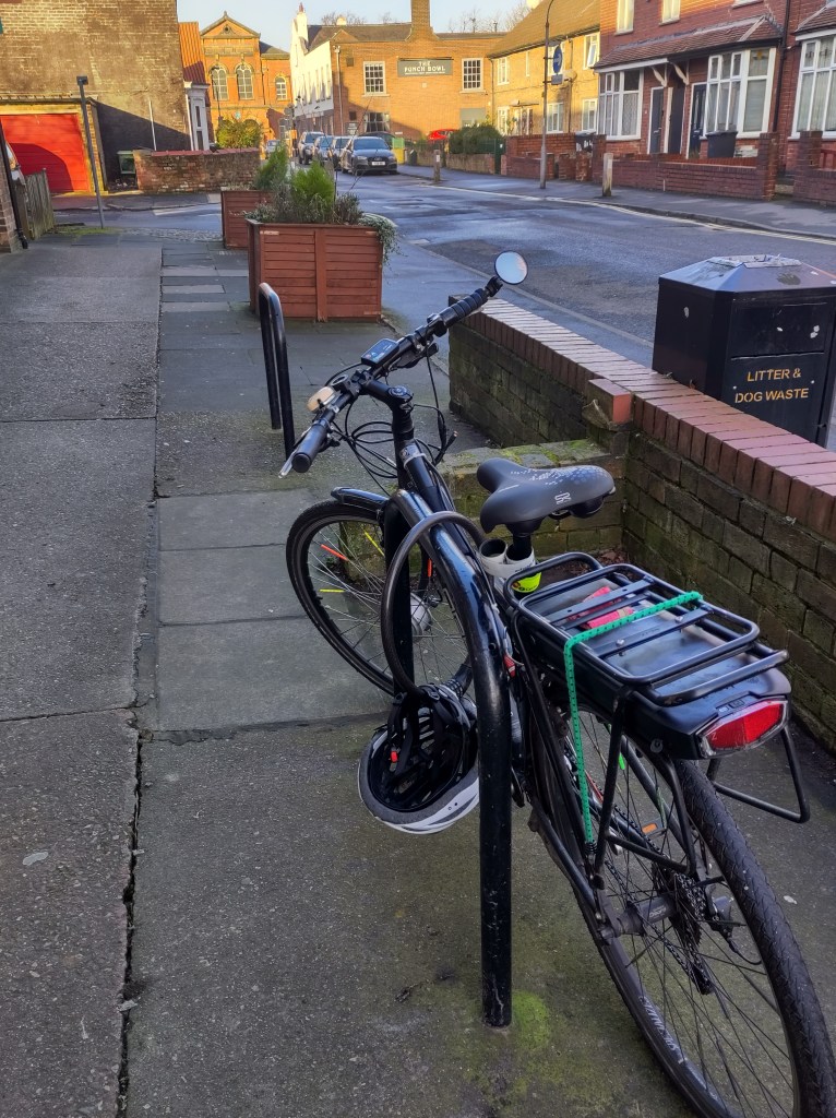 A bicycle secured to a bike rack on a sidewalk, with residential buildings and parked cars in the background.