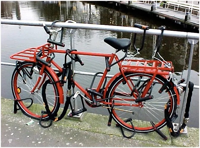 A securely locked red bike with multiple locks attached to a railing near water.