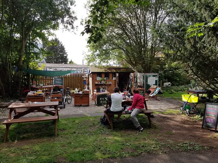 Outdoor scene featuring picnic tables, individuals sitting, and a small refreshment stand surrounded by trees.