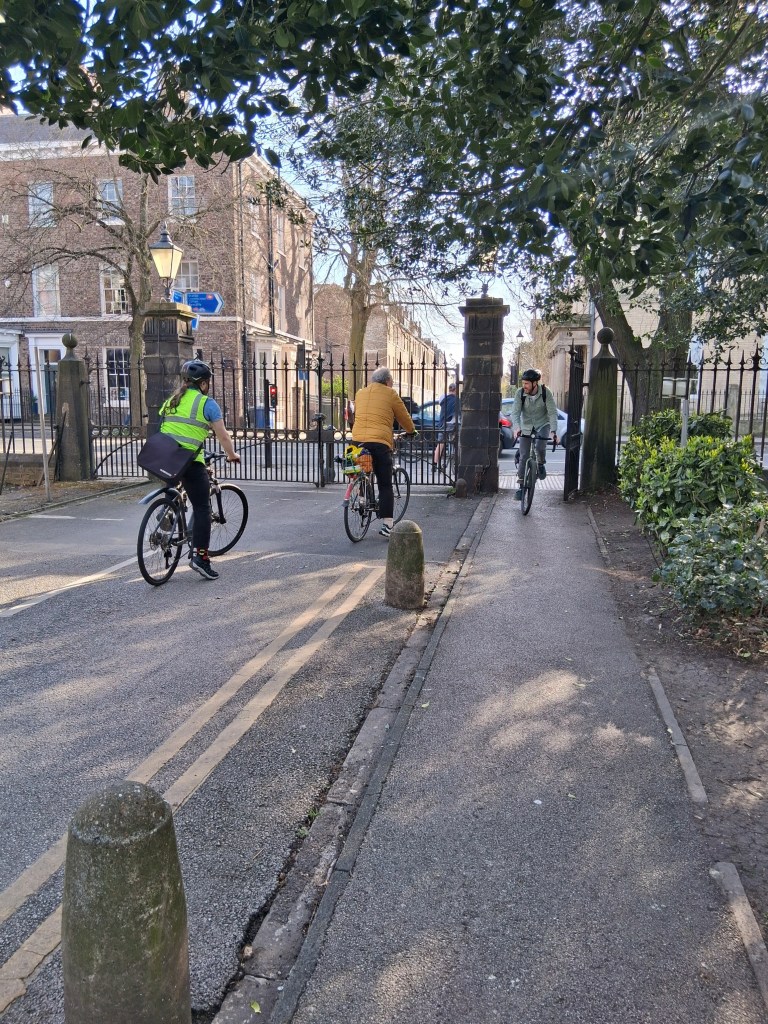 Three cyclists riding along a street near a gated entrance, with trees lining the road and buildings in the background.