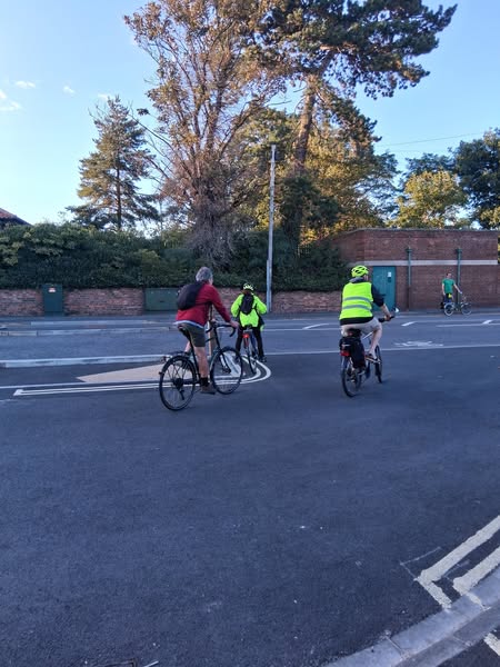 A group of cyclists riding on a street, with trees and a brick wall in the background. One cyclist is wearing a bright yellow high-visibility vest.