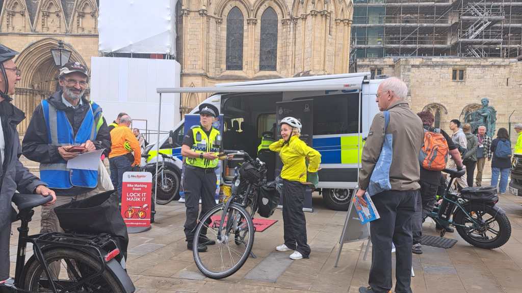 A group of individuals, including police officers and cyclists, gathered near a police bike marking station outside a historic building. The scene includes a police van and signage promoting bike security marking.