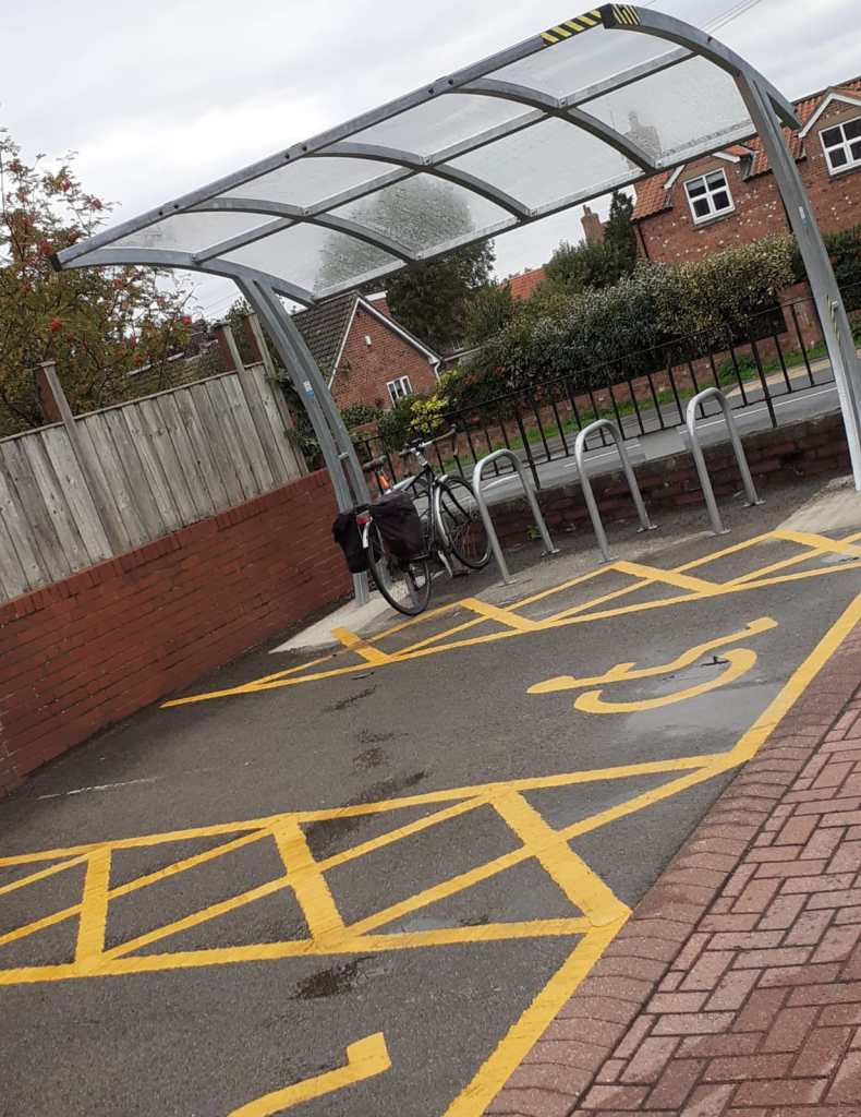 A bike parked under a shelter next to designated bicycle parking spots, surrounded by yellow lines, with residential houses in the background.